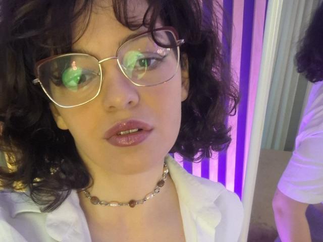 CurlyVixen with curly black hair, wearing glasses, a white shirt, and a silver necklace with brown beads, looking down with slightly parted, shiny pink lips.