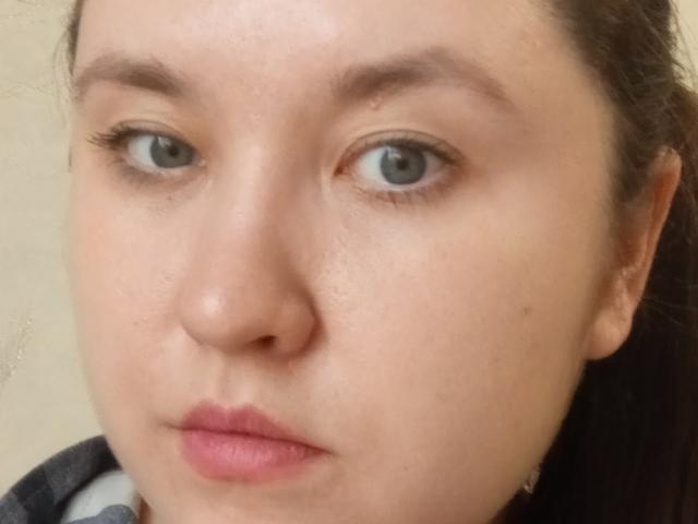 AnnaSova69's face with pale skin, brown eyebrows, green eyes, and pink lips, showing a neutral expression, with dark hair pulled back. Her nose is small and she's wearing a necklace.