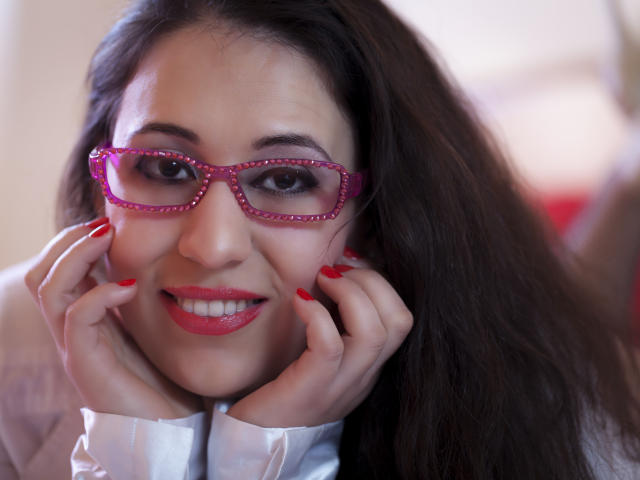 RenattaRosse, com óculos rosa, batom vermelho e unhas vermelhas, sorrindo com as mãos nas bochechas, tem cabelo escuro e está vestindo uma camisa branca. Seus seios não estão visíveis..
