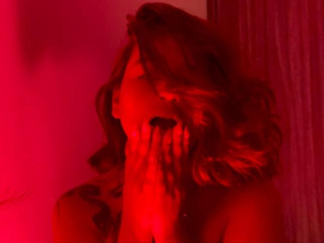 Red-lit photo of SabriinaMorgan with wavy hair, hand covering mouth, visible tattoo on shoulder, nose slightly upturned, red light casting soft shadows.