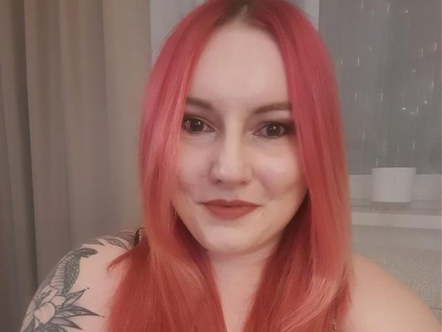 VictoriaDream with pink hair, tattoos on her shoulder, wearing dark lipstick, looking at the camera with a slight smile. Her tits are partially visible.