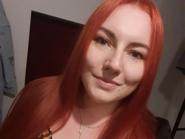 VictoriaDream has red hair, fair skin, and a slight smile with small moles on her chin. She wears a gold necklace, and her brown eyes are relaxed.