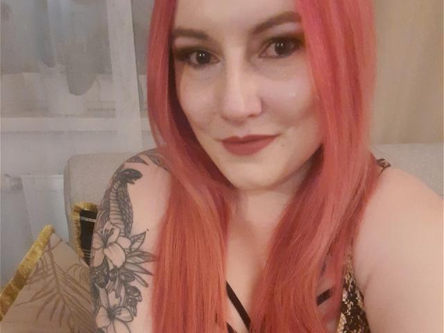 VictoriaDream has pink hair, black eyeliner, and a tattoo on her right shoulder with flowers. She's smiling with red lipstick, wearing a leopard print top, and has small tits.