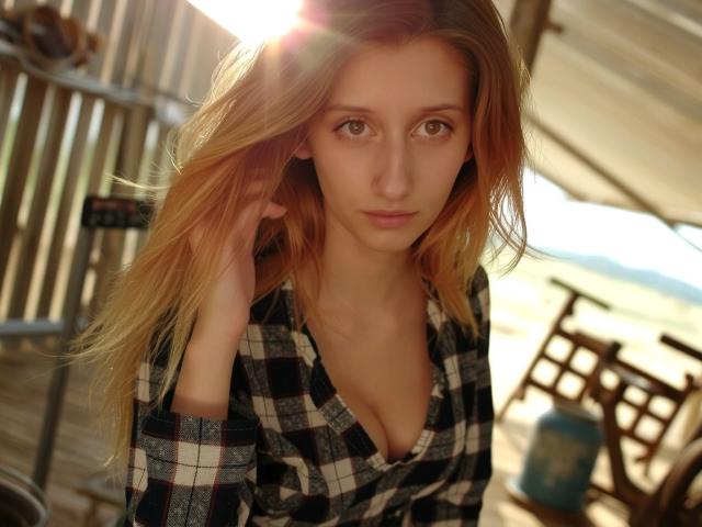 LunaXKiss with long, dirty blonde hair, looking serious, wearing a black-and-white checkered shirt, sun shining on her face, hand in hair, light brown eyes, small tits, wooden background.