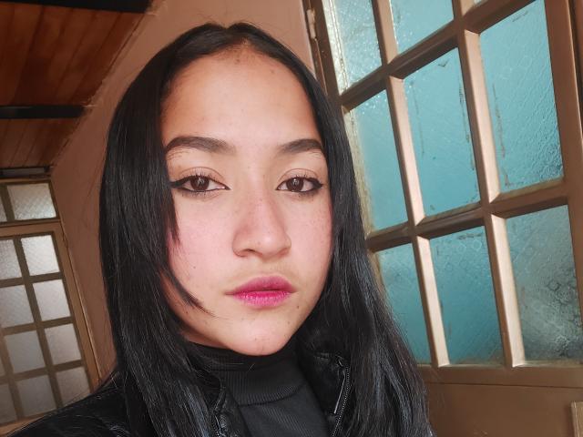 SpringFlower has straight black hair, pink lips, and black eyeliner. She's wearing a black turtleneck and looking at the camera with a neutral expression. Her ass is not visible.