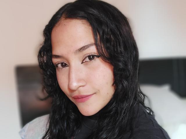 SpringFlower has wavy black hair, brown eyes, and a light skin tone with slight freckles, wearing a black top, and smiling subtly with a slight pink lip.