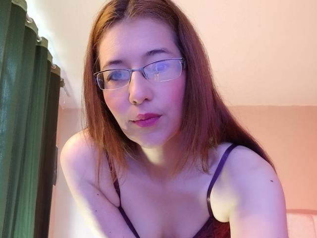 AntonnellaS, avec des lunettes, des lèvres roses et des cheveux bruns, portant un haut violet à bretelles fines, montrant ses seins, légèrement penchée en avant..