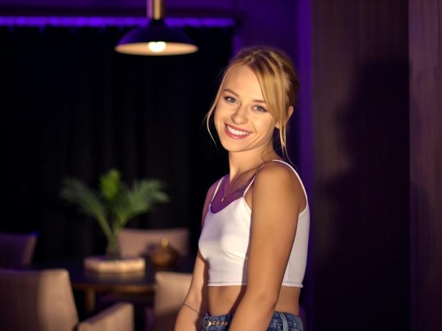 CindyxGlam, blonde girl with small tits, wearing a white crop top and blue jeans, smiling at the camera, standing in a dimly lit room with purple lighting.