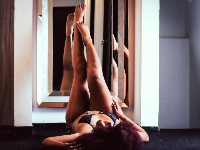 DalilaHeinz is lying on the floor with legs up, wearing black lingerie, reflecting in a mirror. Her dark purple hair is messy, and her tits are visible.