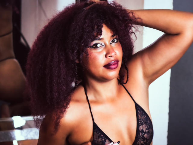 DalilaHeinz with curly dark red hair, wearing a black lace bra, raising one arm, showing her brown skin and purple lipstick, looking at the camera with a confident expression.