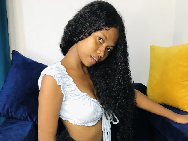 Saloma with long curly black hair, light brown skin, small tits in a blue crop top, sitting on a dark blue and yellow pillow, looking slightly to the side.