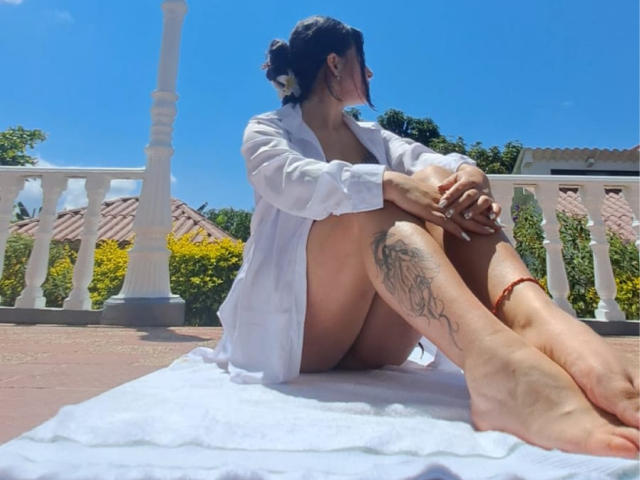 HathorX, naked but for a white shirt, sits on a towel with knees to chest, showing a tattooed leg and red anklet, on a sunny balcony.