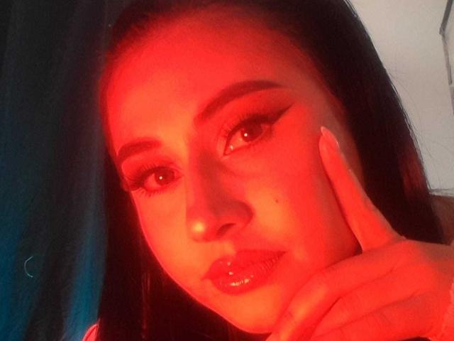 FrancheskaaWaalsh's red-lit face, dark hair, and glossy lips, with her finger near her cheek, showing off long, thick, and curled eyelashes. Her nipples are not visible.