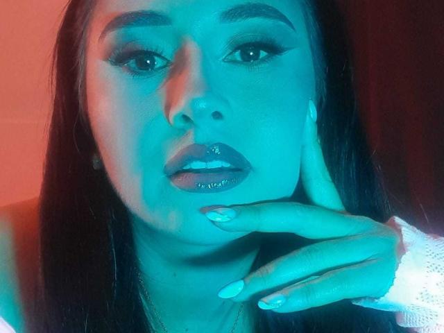 Francheska Walsh, con labios morados brillantes, rostro iluminado en azul, cabello largo y oscuro, y uñas cuidadas, tocándose la barbilla, vestida con una camisa blanca de manga larga..