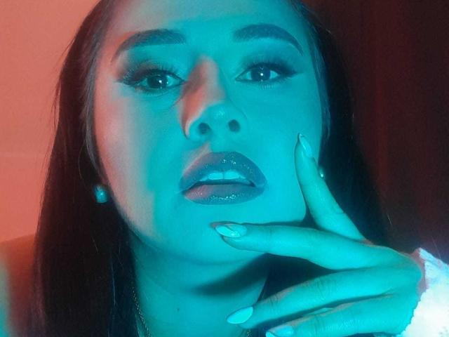 FrancheskaWaalsh has blue-tinted skin, glossy purple lips, long dark hair, and is touching her chin with blue-manicured nails, wearing pearl earrings.