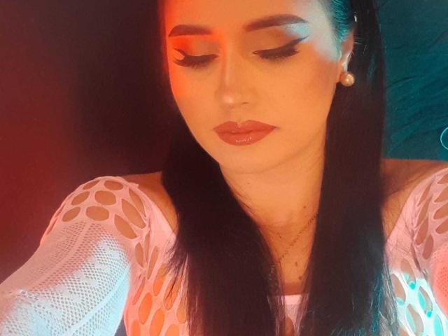 FrancheskaWaalsh with long black hair, wearing a white mesh top with holes, has red-orange and teal lighting on her face, and is looking down with shiny red lips.