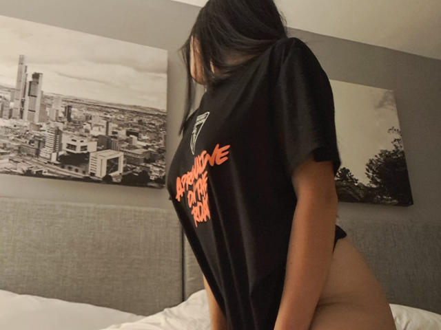 HathorX with long black hair, wearing a black T-shirt, exposing bare ass, in a gray-walled bedroom with black-and-white cityscape paintings on the wall.