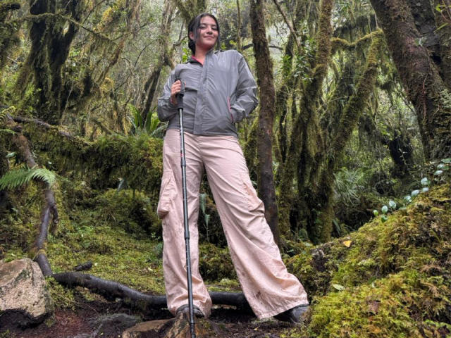 HathorX with medium tits, standing in a mossy forest, wearing a gray jacket, pink pants, and holding a hiking pole, smiling confidently.