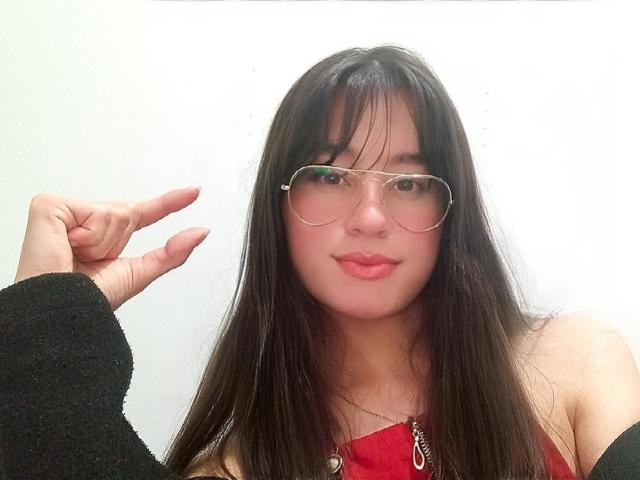 MaddieHottie with long black hair, wearing gold aviator glasses, a red top, black sleeves, and a necklace, pointing with her right hand, smiling with pink lipstick.