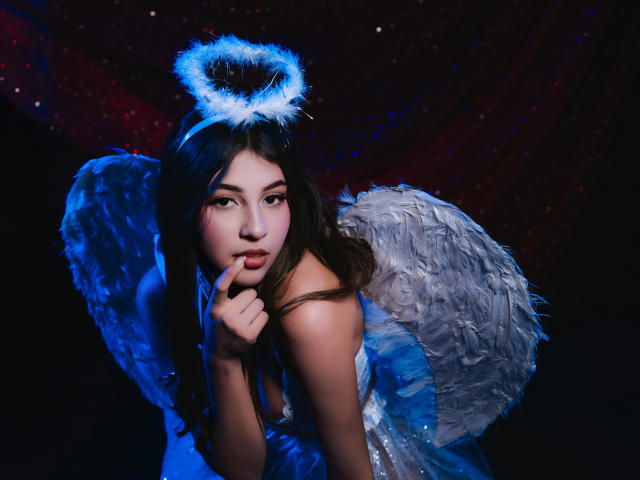 MillyLane with a halo and white feather wings, her finger touching her pink lips, looking seductively at the camera, her brown hair and blue dress contrasting.