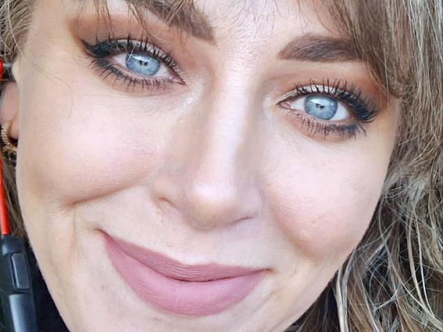 El rostro de CandyLadyZ, con sus ojos azules y una ligera sonrisa con labios rosados, delineador de ojos oscuro y cejas marrones. Tiene cabello rubio, ligeras arrugas y una nariz rosada. Está sosteniendo un objeto rojo y negro..