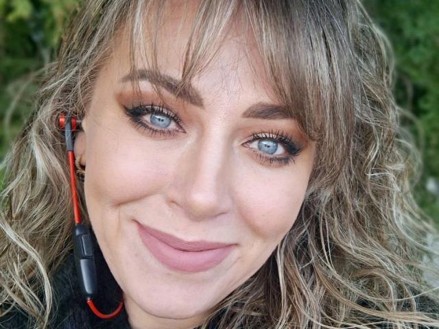 CandyLadyZ has wavy blonde hair, blue eyes, and a cute smile. She's wearing earbuds with red and black cords, and has subtle makeup with black eyeliner.