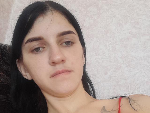 CaseyJeannie has pale skin, black hair, brown eyes, and a small pimple on her left cheek. She's wearing a red top with a visible tattoo on her right shoulder.