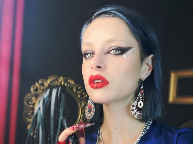 GoddessDivonix met blauw haar, witte make-up stipjes, rode lipstick, een doorboorde lip, rode nagels, een blauwe jurk, zilveren oorbellen, kijkt omhoog, hand op kin, donkere achtergrond, elegant..