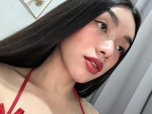 PickmeTrinaa has long black hair, red lipstick, and is wearing a red halter top. Her eyebrows are thick and she has a slight smile on her face.