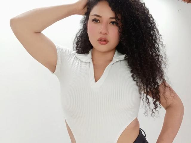 CarlaMuille with curly black hair, wearing a white ribbed V-neck top, exposing her side, with her hand in her hair, looking at the camera with pink lips.