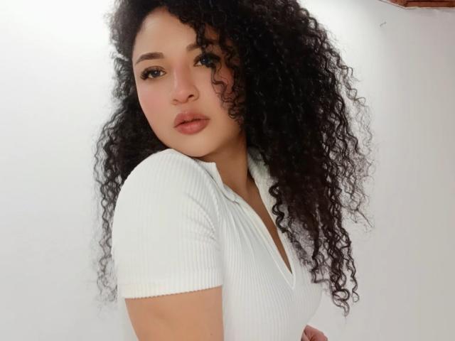 CarlaMuille with curly black hair, white ribbed top, and pouty pink lips, looking at camera with a slight turn, showing her tits in a relaxed pose.