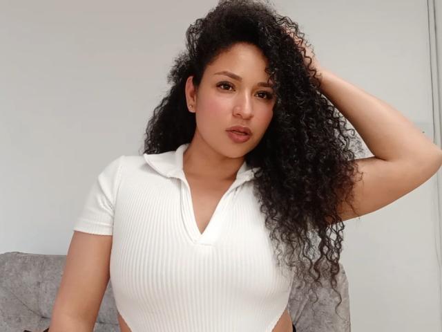CarlaMuille mit lockigem, schwarzem Haar, trägt ein weißes, geripptes Crop-Top, das ihre Brüste freilegt. Sie hat eine Hand in ihrem Haar und blickt mit leicht gerunzelter Miene in die Kamera..