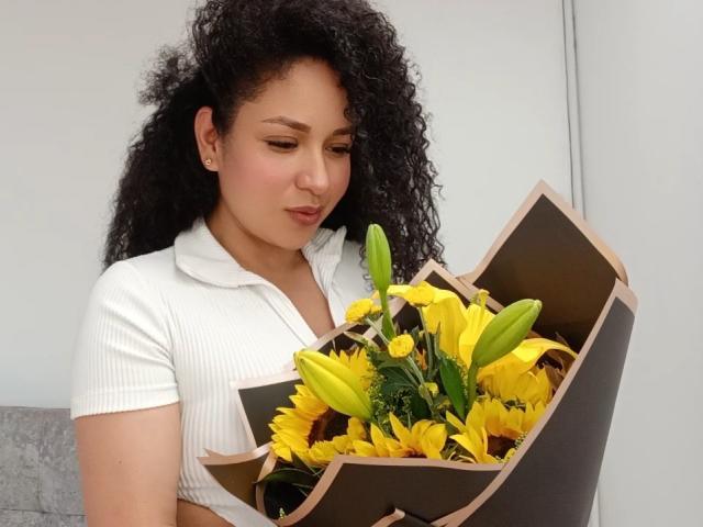 CarlaMuille with curly black hair, white ribbed shirt, holding bouquet of yellow lilies and sunflowers, looking down, brown paper wrapping, cute, soft smile, subtle makeup, casual setting.