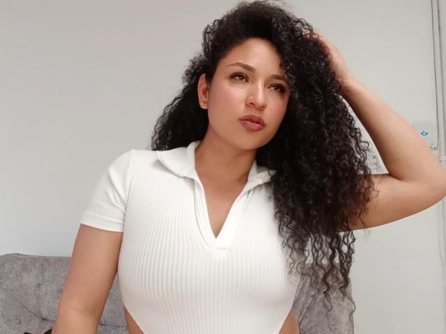 CarlaMuille, con cabello negro rizado, vestida con un polo blanco acanalado, con la mano en el pelo, mirando a la cámara, mostrando sus pechos, piel marrón clara, maquillaje sutil, labios fruncidos, sentada..