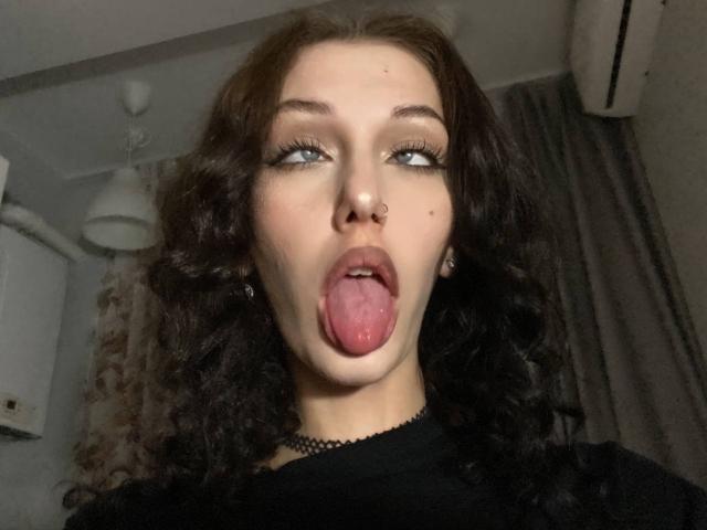 LexxiBlue has curly brown hair, blue eyes, and a nose ring. She's sticking out her pink tongue with her mouth open, wearing a black choker and dark top.