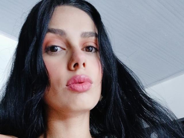 Anais Hilton tiene el cabello negro, lápiz labial rosa y ojos marrones, con una expresión ligeramente seria, mirando directamente a la cámara. Su nariz es pequeña y su piel es clara..