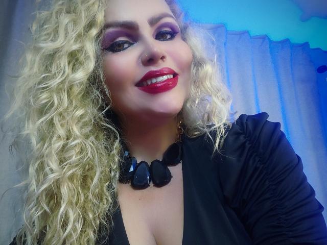 AnnaRichie with curly blonde hair, red lipstick, black necklace, and black top, smiling at the camera, showing her boobs slightly in a low-cut top.