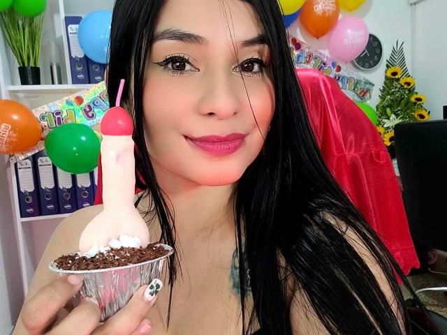 AliceNova with long black hair, pink lipstick, holding a chocolate cupcake with a dildo on top, pink straw, chocolate crumbs, and white nail polish, smiling.