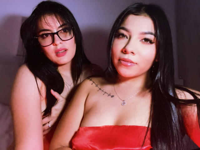 KymAndNala with long black hair, glasses, and red strapless top, showing her tits, standing next to KymAndNala with similar hair, looking at camera, no clothes.