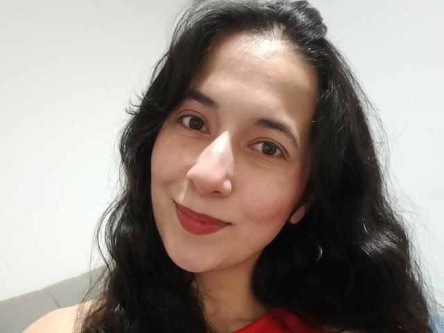 AnneStone has wavy black hair, brown eyes, and red lipstick. She's smiling with a slight blush on her cheeks. Her red top is visible. She's attractive.