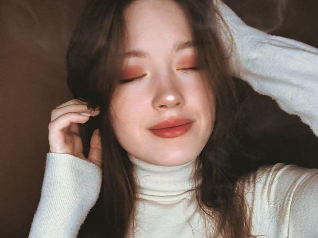 StarBreaker has wavy brown hair, closed eyes with pinkish-orange eyeshadow, and a slight smile. She's wearing a white turtleneck, with one hand touching her head.
