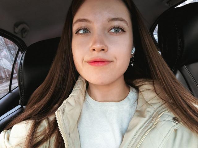 StarBreaker has long brown hair, green eyes, and a slight smile. She's wearing a white shirt and beige jacket with black leather car seats in the background.