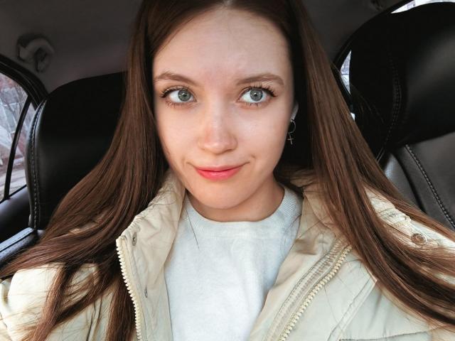 StarBreaker has green eyes, long brown hair, and a cute smile. She's wearing a beige jacket over a white shirt, sitting in a car with black leather seats.
