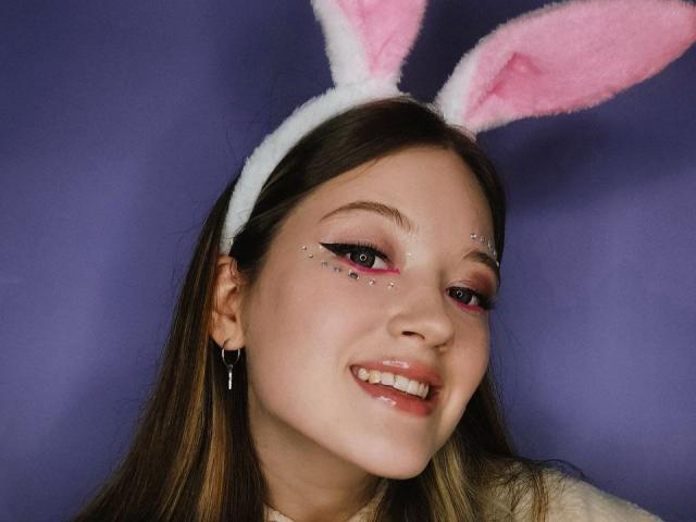 StarBreaker with pink and white fuzzy bunny ears, smiling, has glitter on her eyelids, pink eyeshadow, white headband, long brown hair, and is wearing a cross earring.