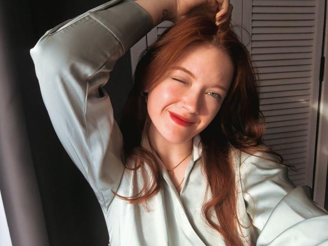StarBreaker with red hair, wearing a light blue shirt, smiling with one eye closed, raising arm to touch head, showing a tattoo on her wrist.