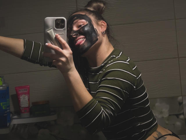JudyGrace with black mask, green striped shirt, tongue out, taking selfie in mirror, showing tattoos, and holding silver iPhone. Her hair is in a bun.