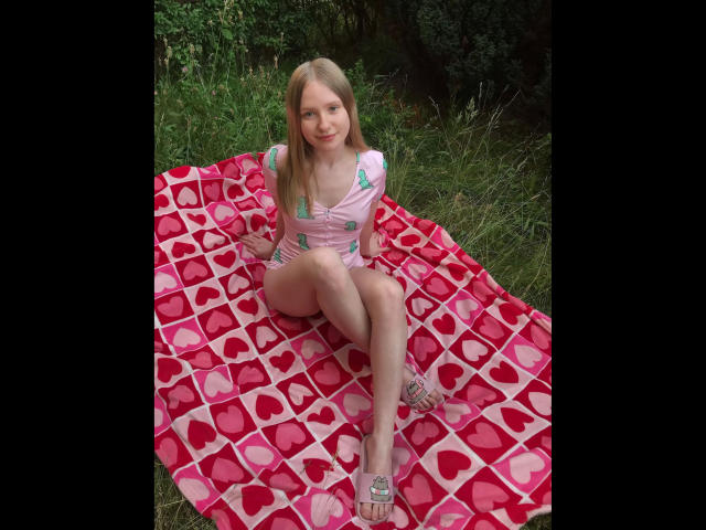 MissNancy-hot with small tits, sitting on a red heart-patterned blanket, wearing a pink top, green shorts, and silver sandals, in a grassy outdoor area.