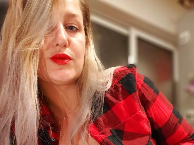MayaTrainor with blonde hair, red lipstick, and red-black checkered shirt, looking at camera, slightly wavy hair covering one eye, subtle smile, soft lighting, casual selfie.