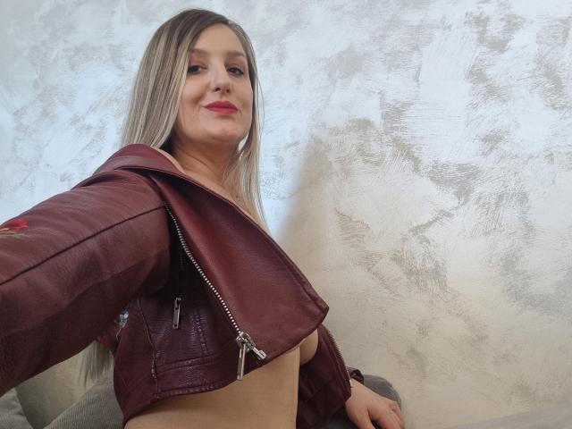 Maya Trainor mit langen blonden Haaren, einer roten Lederjacke und freiliegenden Brüsten, lächelt in die Kamera. Der Reißverschluss ihrer Jacke ist teilweise geöffnet, wodurch ihre nackte Brust sichtbar ist..