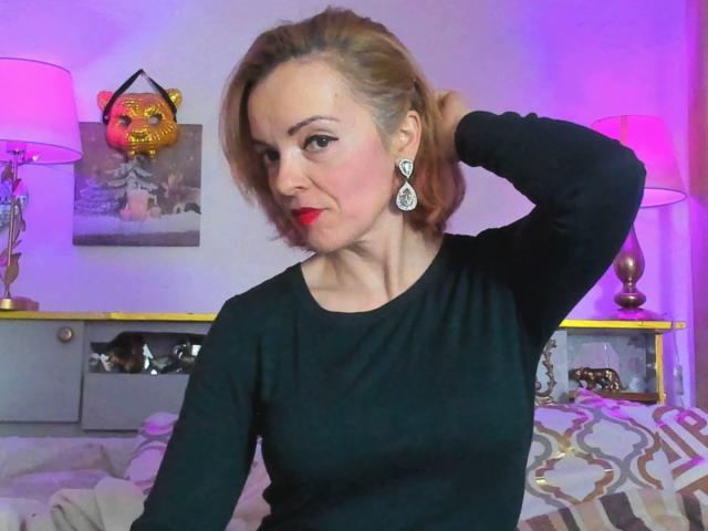KateRouge with red lipstick, black long-sleeve shirt, and teardrop earrings, hand in hair, looking at camera, pink and purple room, lamp, decorative lion, bed with patterned cover.